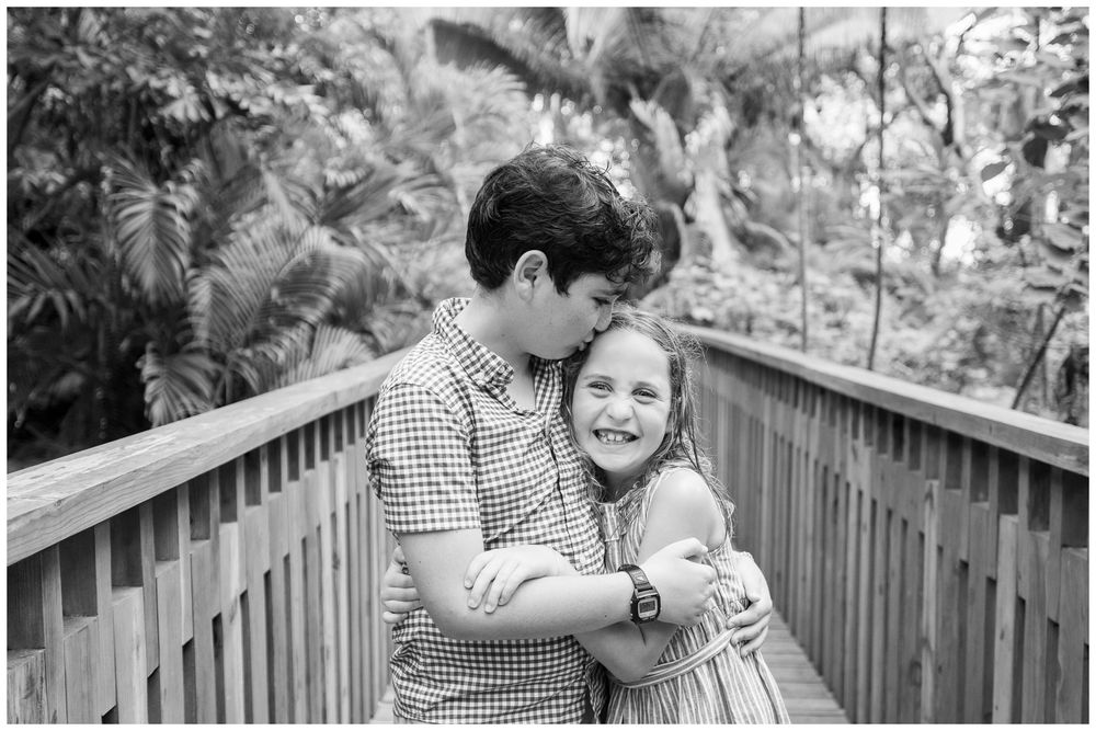 Black and white photo of a boy kissing a laughing girl on a wooden bridge surrounded by lush greenery.