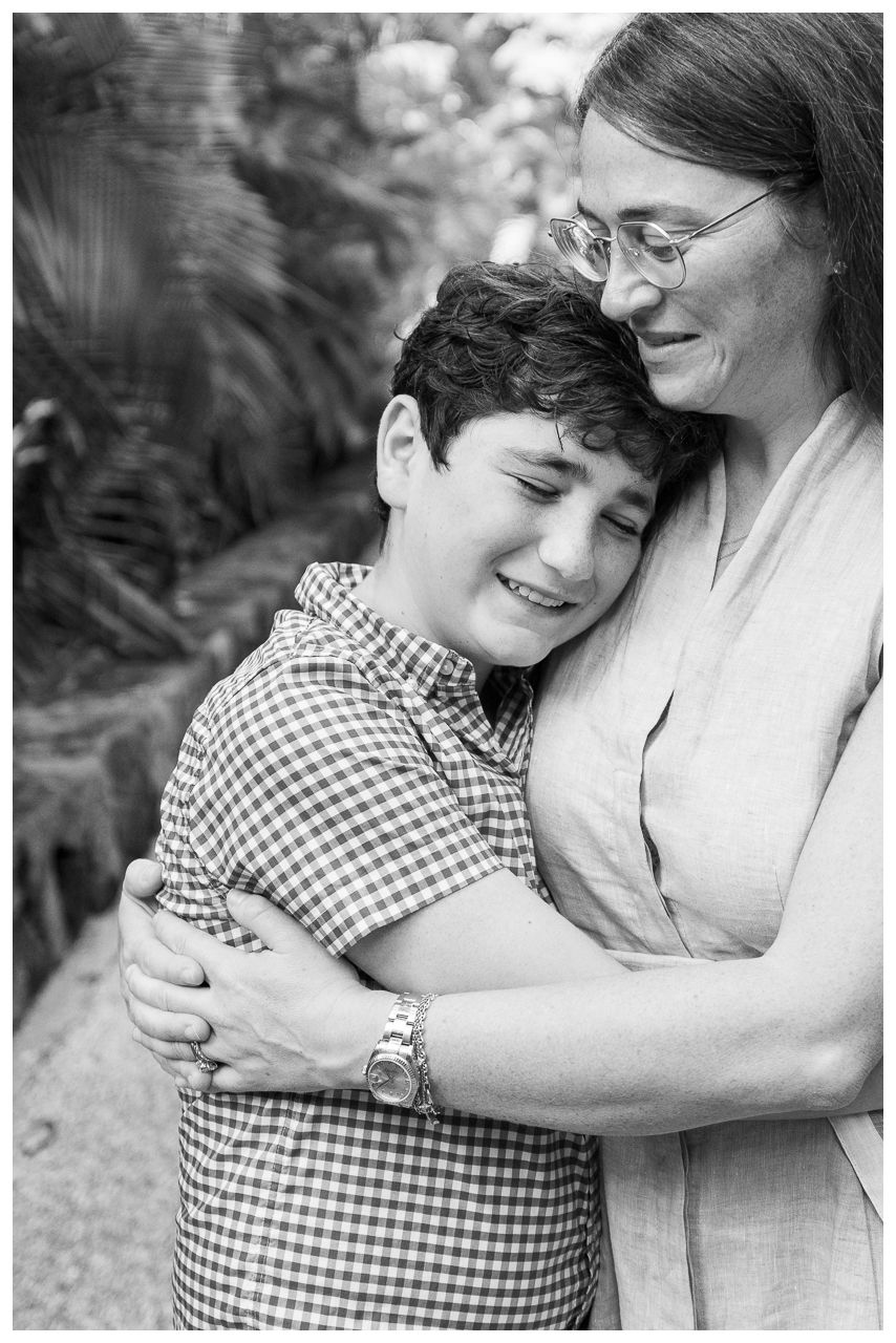 Black and white photo of a boy hugging a woman wearing glasses, both smiling warmly outdoors.