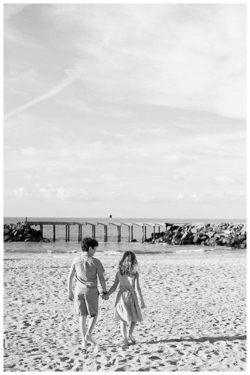 Black and white photo of a couple holding hands walking on a sandy beach toward a pier and rocky jetty.
