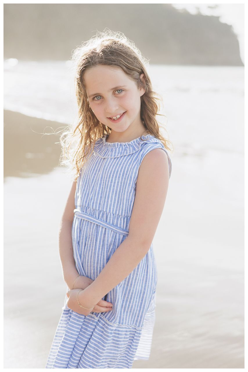 A young girl in a blue striped dress smiles on a beach, her hair blowing gently in the breeze.
