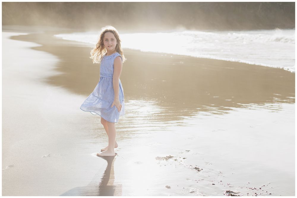 A young girl in a blue dress walks barefoot along a sunlit beach shoreline with gentle waves.