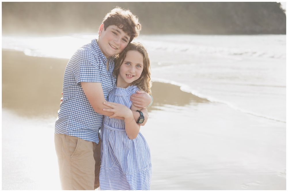 Two siblings hugging and smiling on a sunlit beach, boy in blue gingham shirt and girl in blue dress.