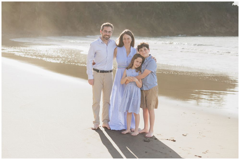 Family of four posing together on a sunny beach, parents and two children smiling in coordinated blue and tan outfits.