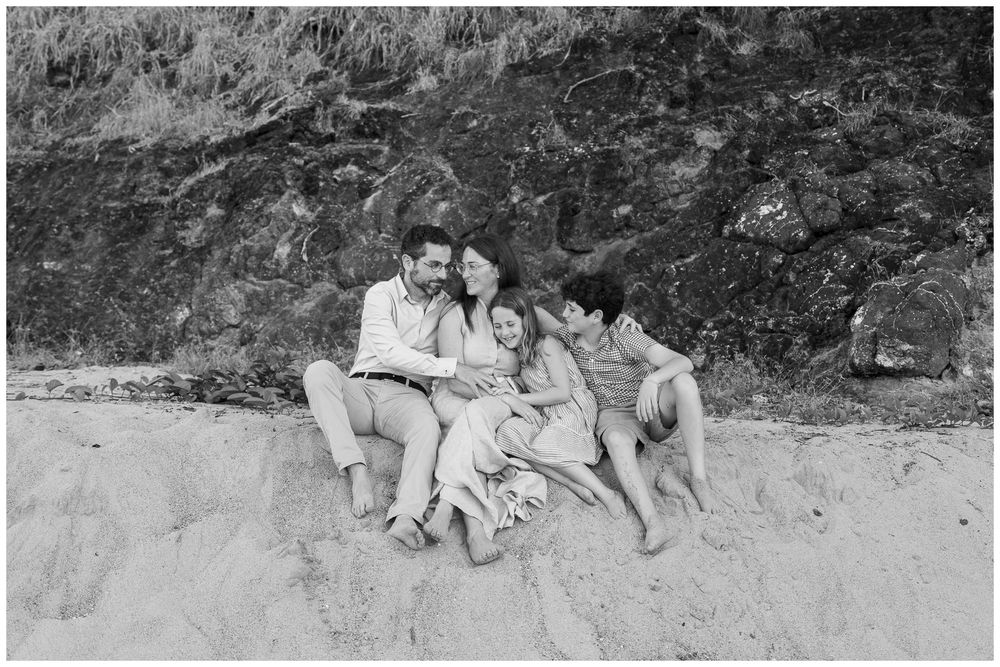 Black and white photo of a family of four sitting together on a sandy beach with rocky cliffs behind them.
