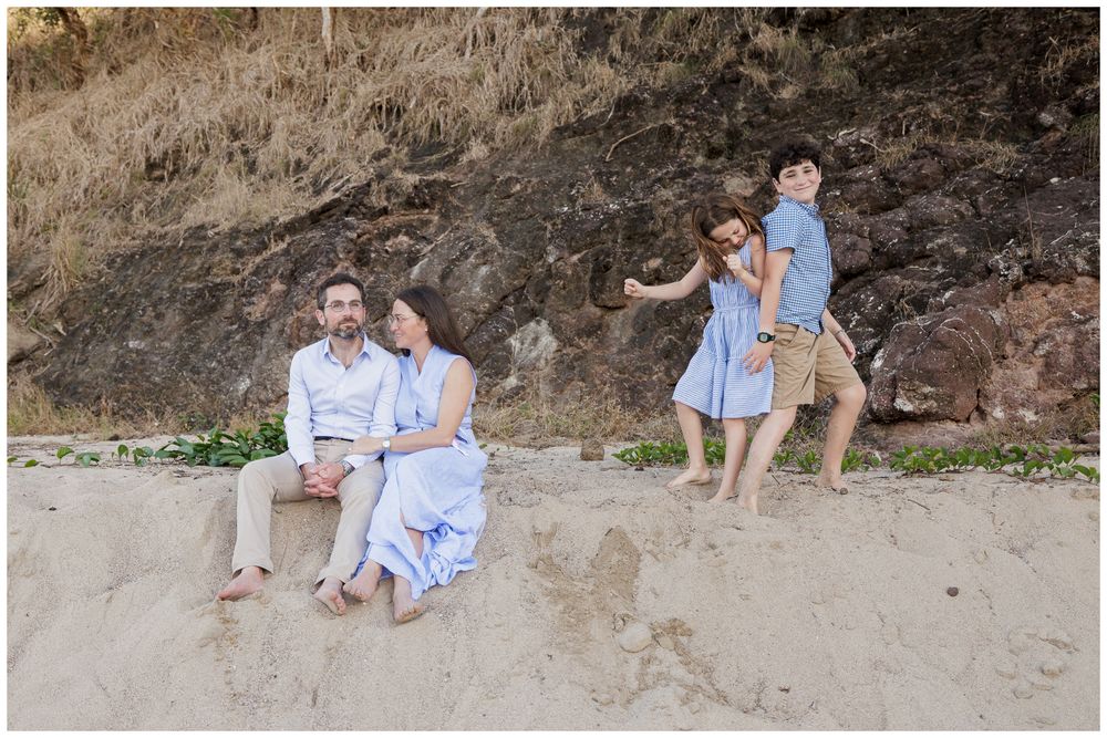 Family beach portrait with parents sitting on sand while children play near rocky cliff backdrop.