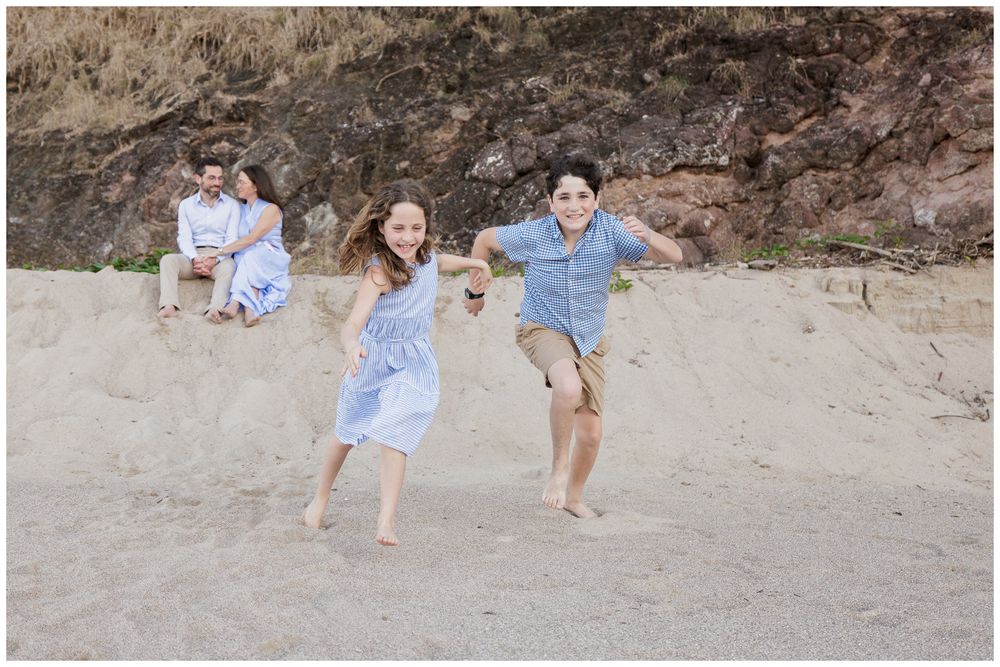 Two children running toward camera on sandy beach while parents sit together in background.
