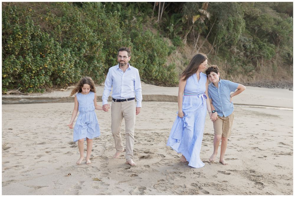Family of four walking on sandy beach in coordinating blue outfits, with lush green trees in the background.