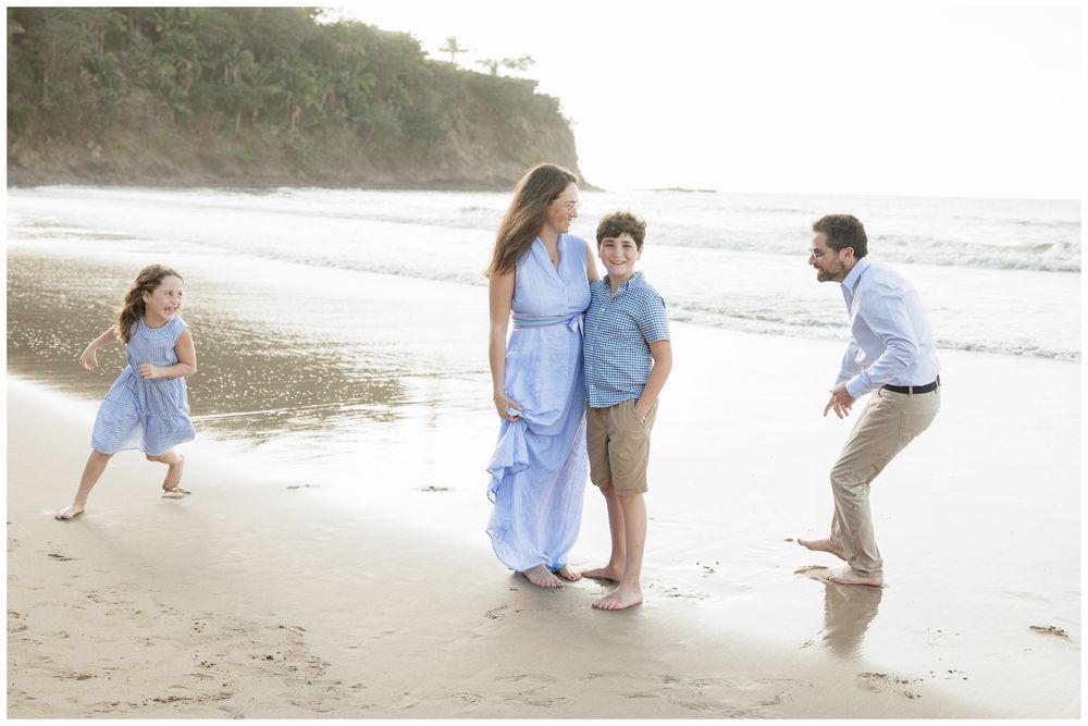 Joyful family of four playing on a tropical beach, children running while parents smile in matching blue outfits.