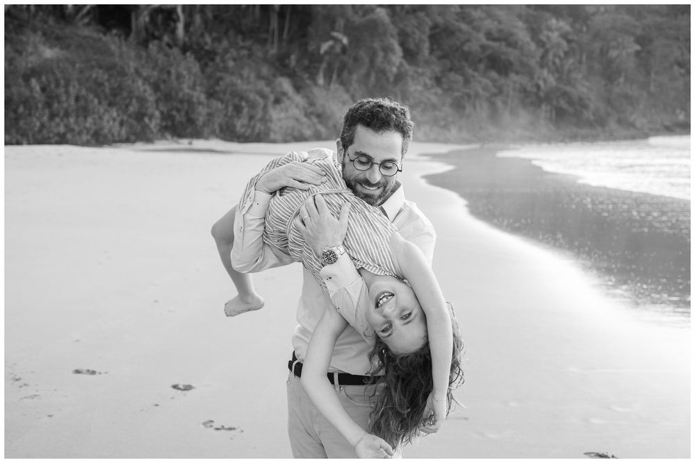 Black and white photo of a laughing couple playing on a sandy beach, man lifting woman over his shoulder.