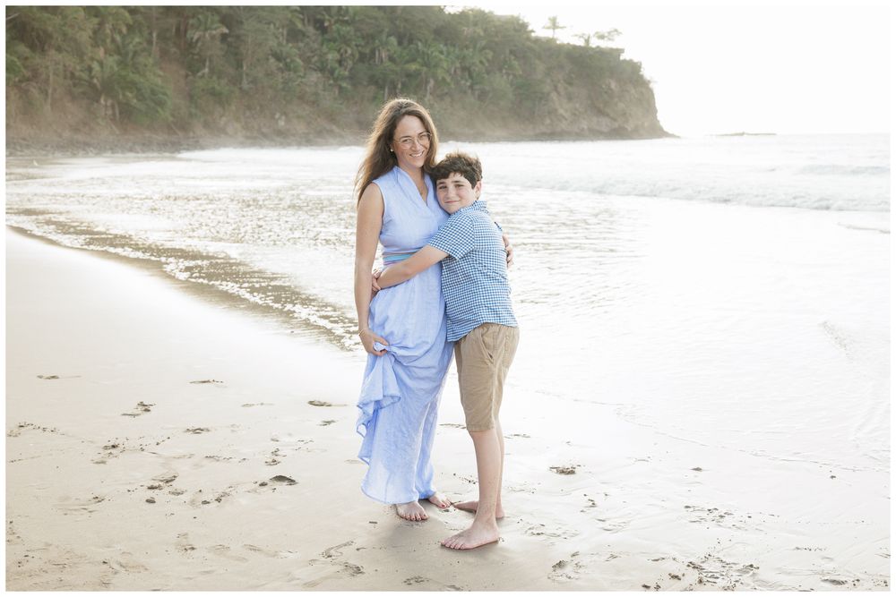 A mother and son share a warm embrace on a scenic beach, she wearing a flowing blue dress.
