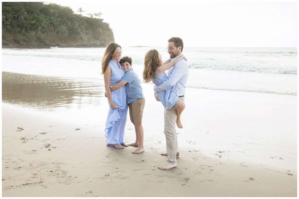 Family of four posing together on a sandy beach at sunset, wearing coordinating light blue outfits.