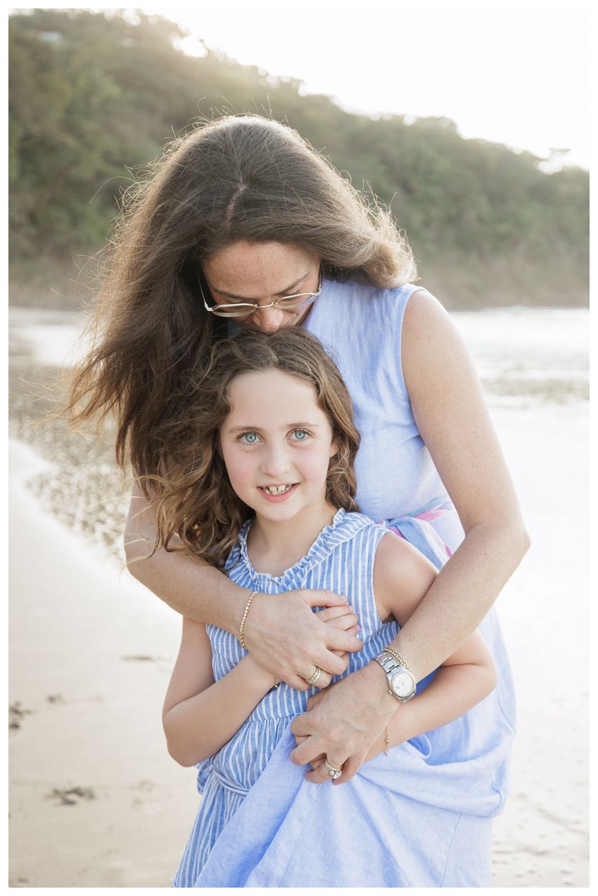 A mother hugs her smiling daughter on a beach, both wearing blue dresses in a warm, golden-light portrait.