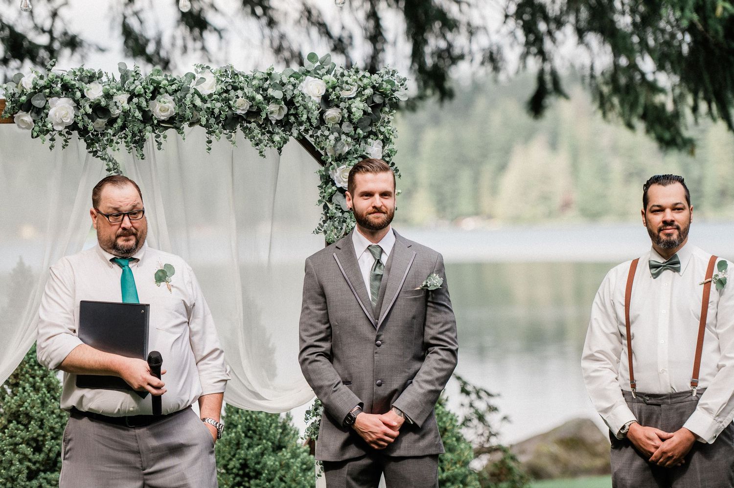 Three men stand at an outdoor wedding ceremony altar decorated with white flowers and greenery near a serene lake.