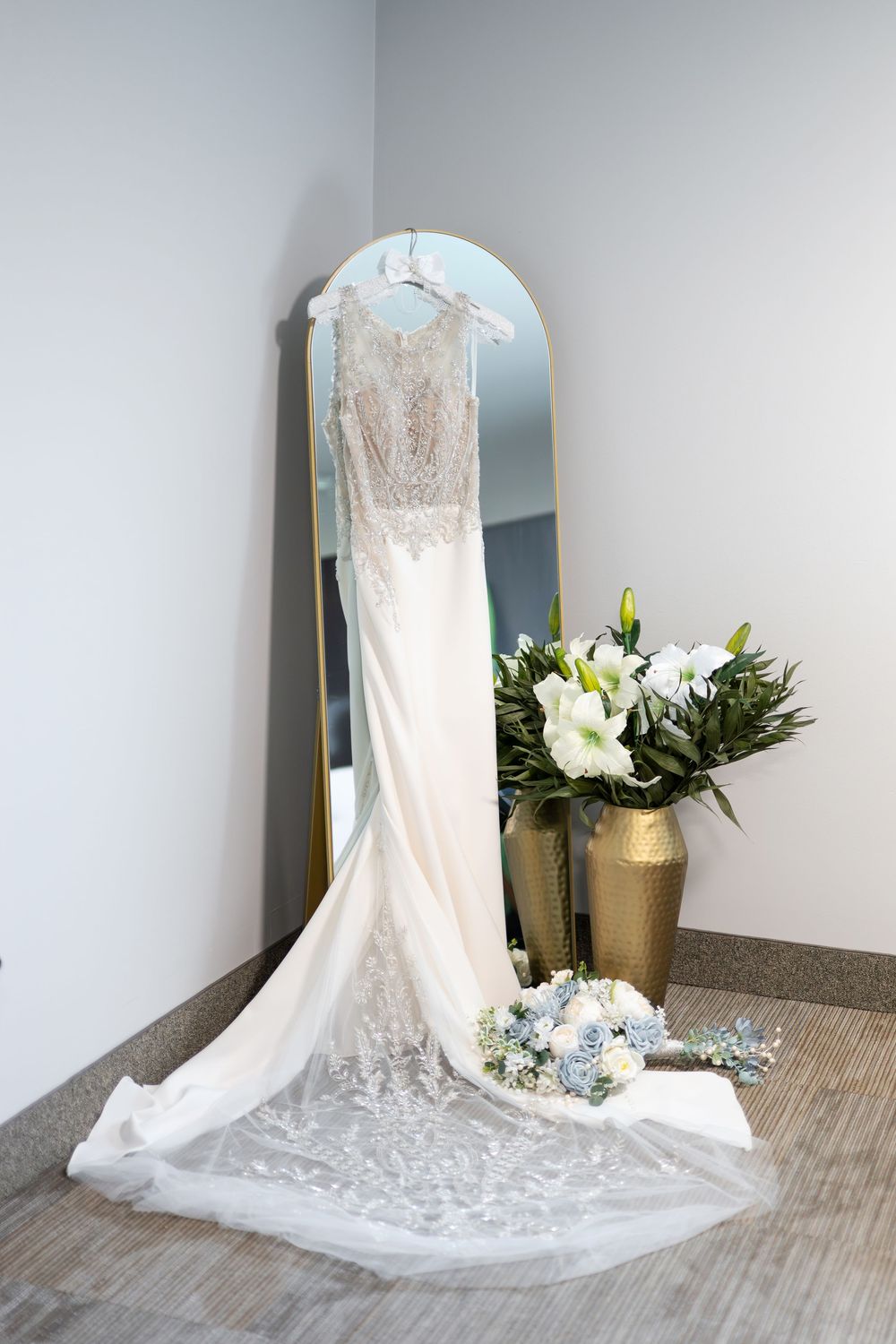 Elegant lace wedding gown hangs on mirror beside gold vase with white lilies and bridal bouquet.