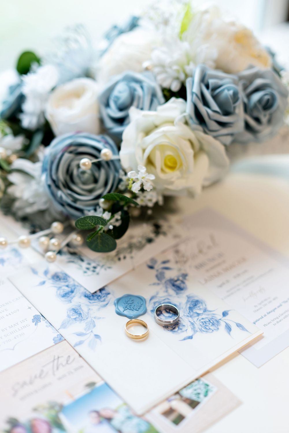 Wedding rings rest on floral blue stationery beside a bouquet of dusty blue and white roses.