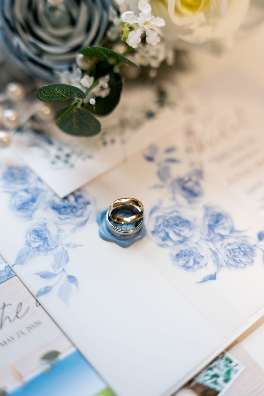 Two wedding bands rest on blue floral wedding stationery beside a bouquet of roses and greenery.