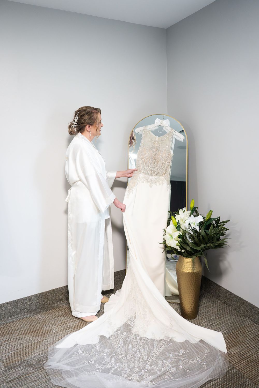 Bride in white robe admires her wedding gown displayed on mirror before ceremony.