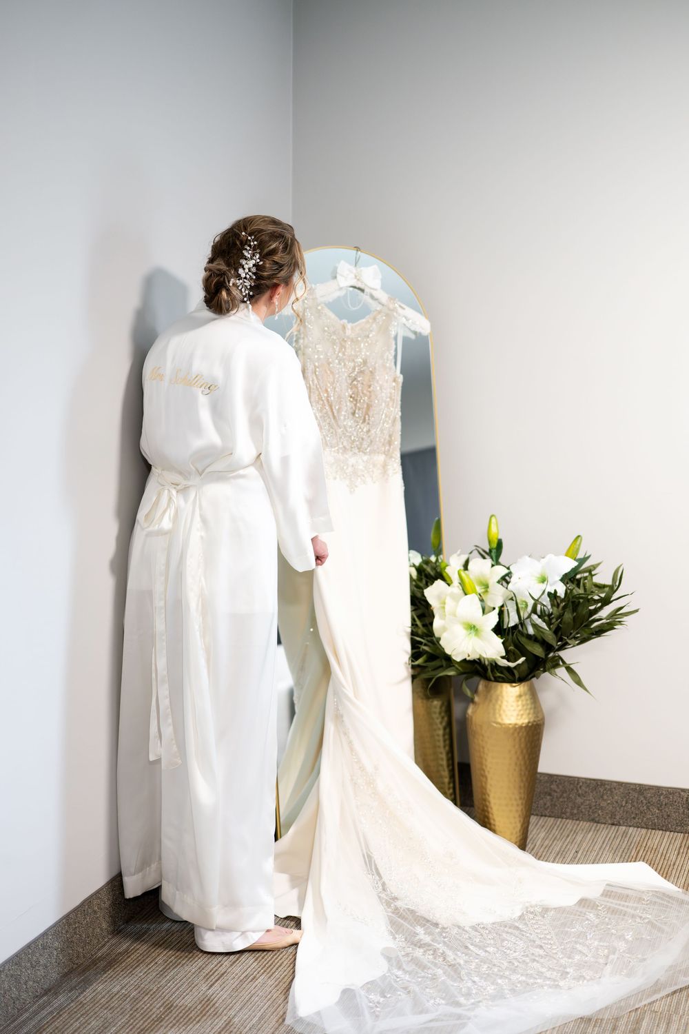 Bride in white robe viewing her lace wedding gown on a mirror beside white floral arrangement.