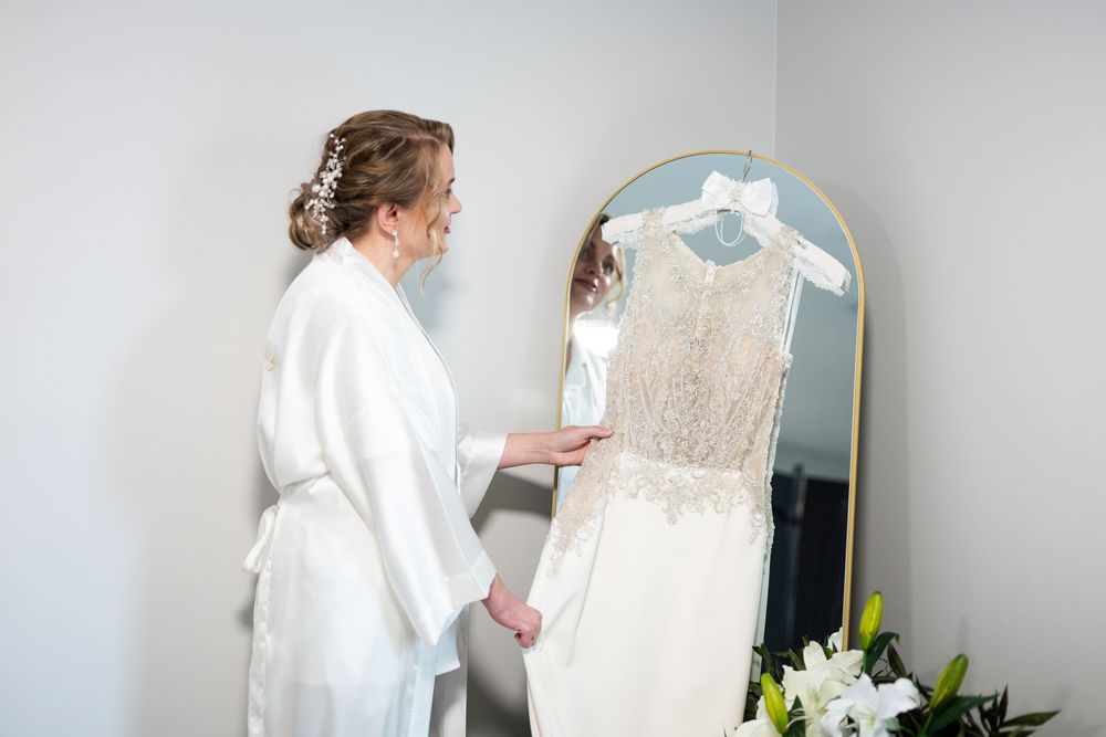 Bride in white robe admires her lace wedding dress hanging on a gold mirror before the ceremony.