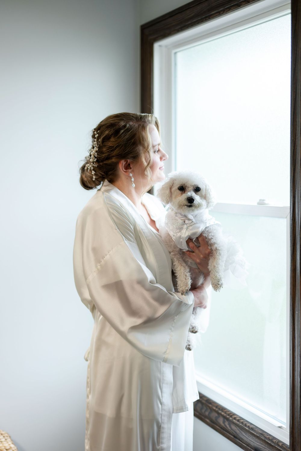 Bride in white robe holding a fluffy white dog while gazing out a window during wedding preparations.