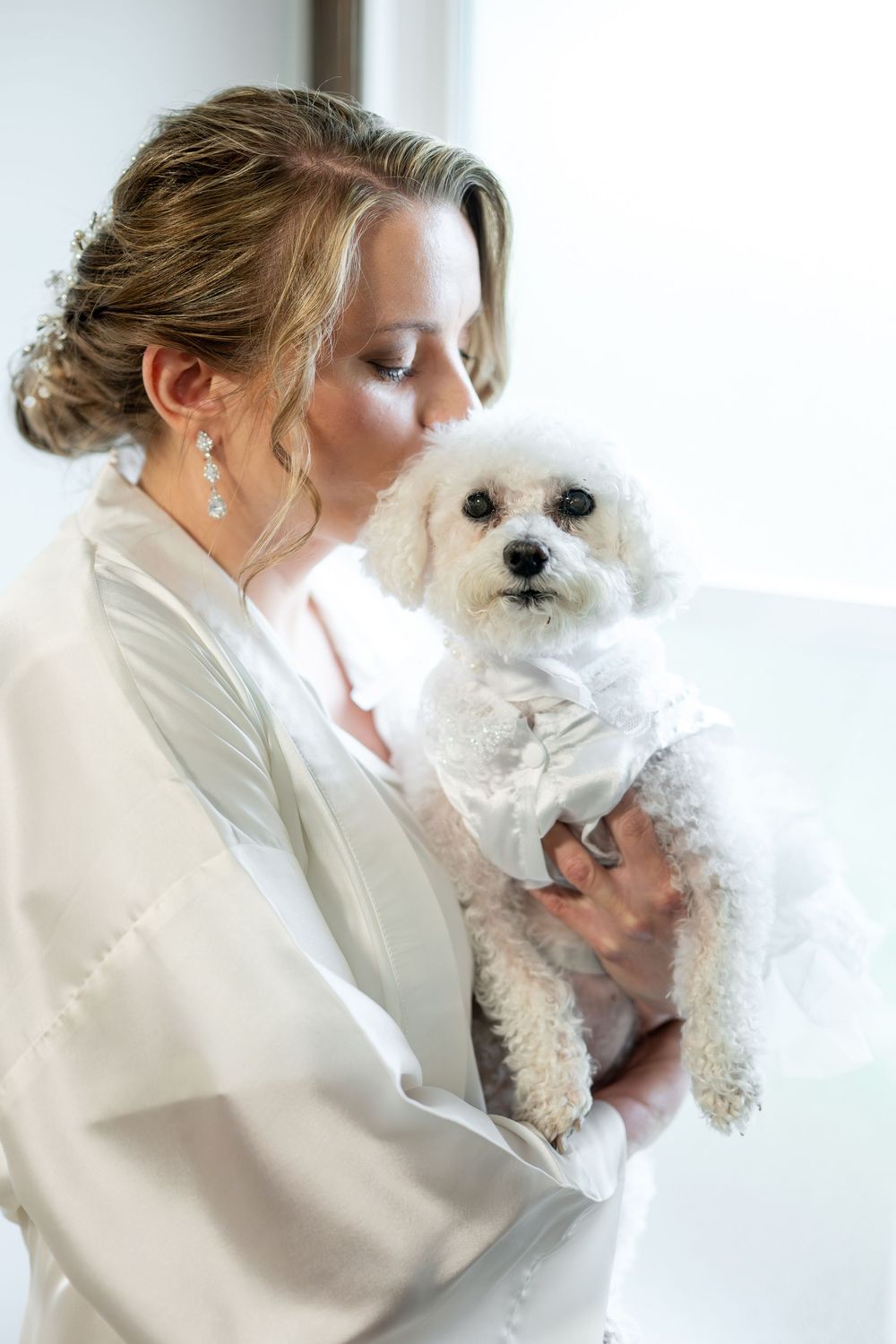 Bride in a white robe kisses her small white Bichon Frise dog dressed in a bow tie.
