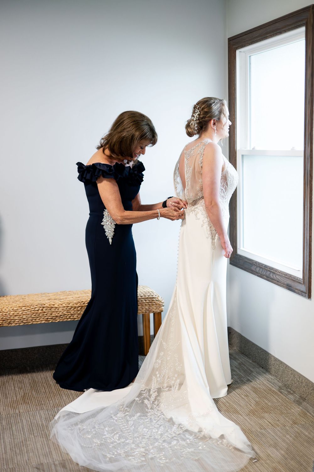 Mother of the bride fastens the back of the bride's lace wedding gown near a window.