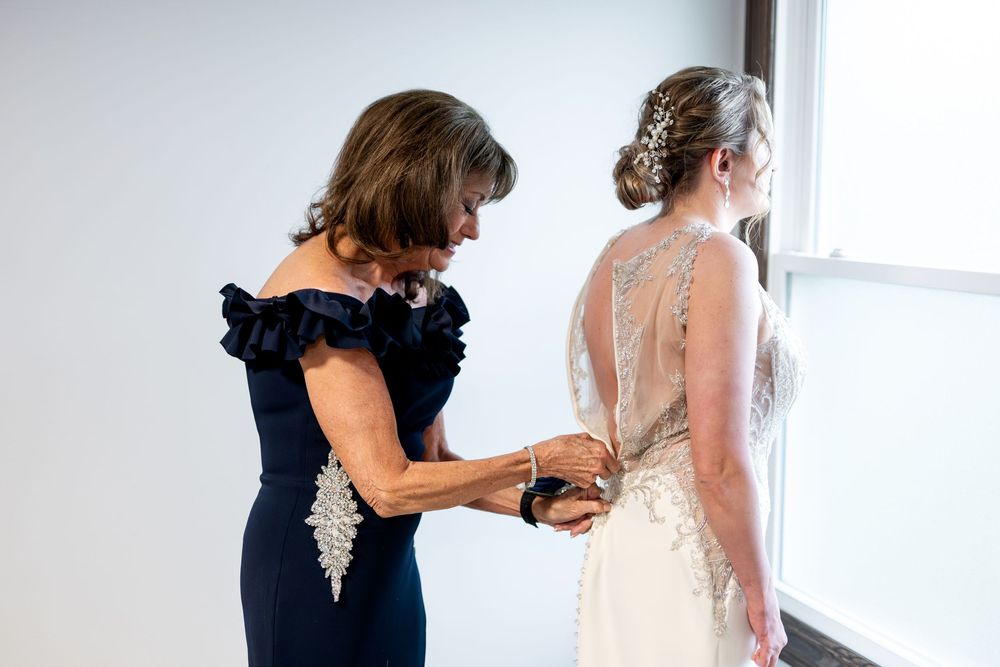 Mother helping bride fasten the back of her beaded lace wedding gown near a bright window.
