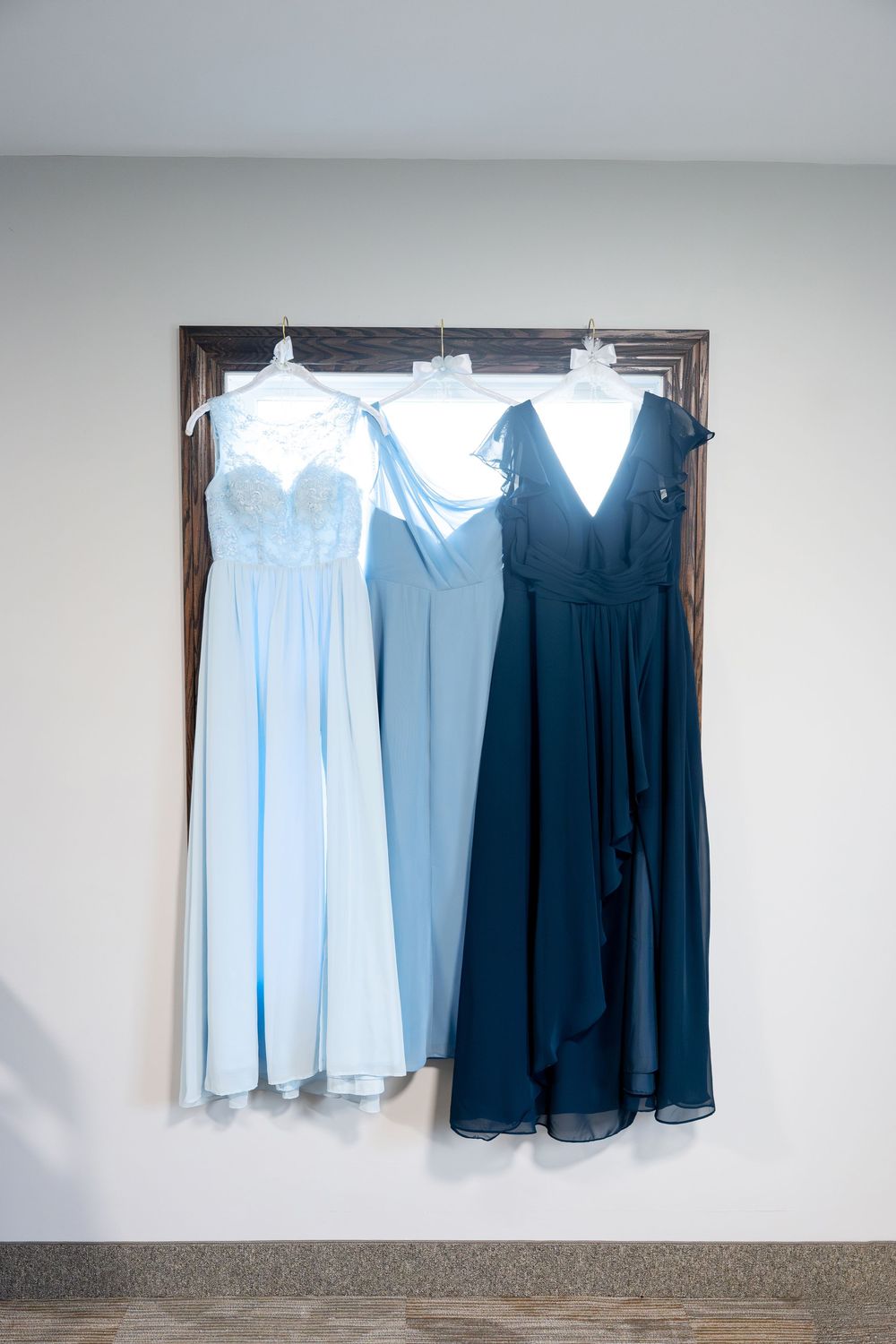 Three bridesmaid dresses in light blue, sky blue, and navy hanging on a rustic wooden frame.