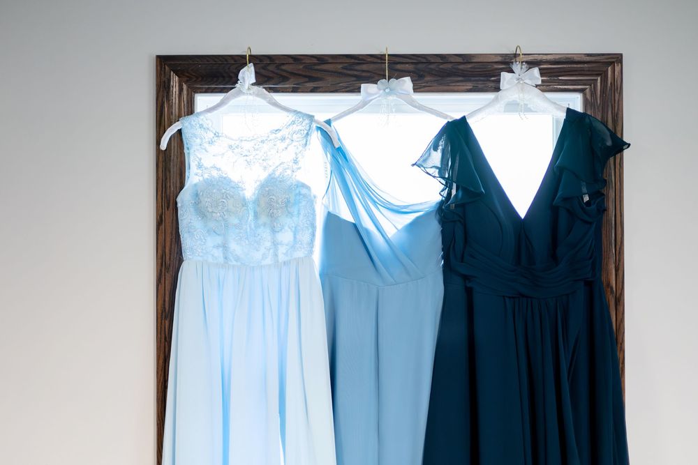 Three bridesmaid dresses in light blue, medium blue, and dark navy displayed on a wooden hanger frame.