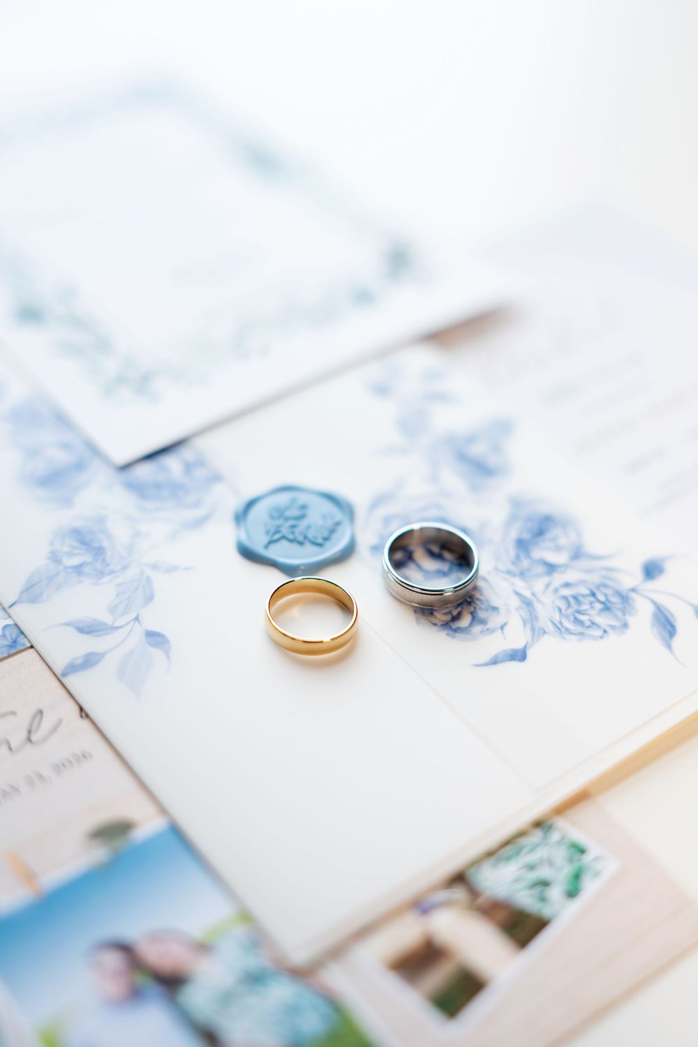 Two wedding bands, one gold and one silver, resting on blue floral wedding invitation with wax seal.
