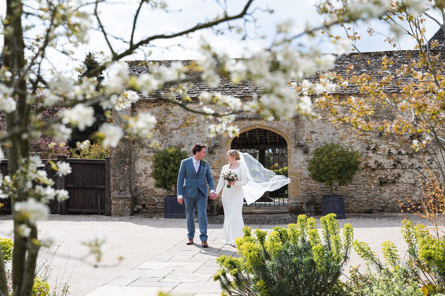 Bride and groom walking hand in hand in a sunny courtyard framed by white blossoms at a rustic stone venue.