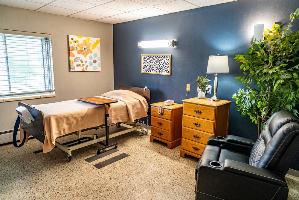 A cozy skilled nursing room featuring a bed, wooden dresser, lamp, recliner chair, and blue accent wall with artwork. branding photography