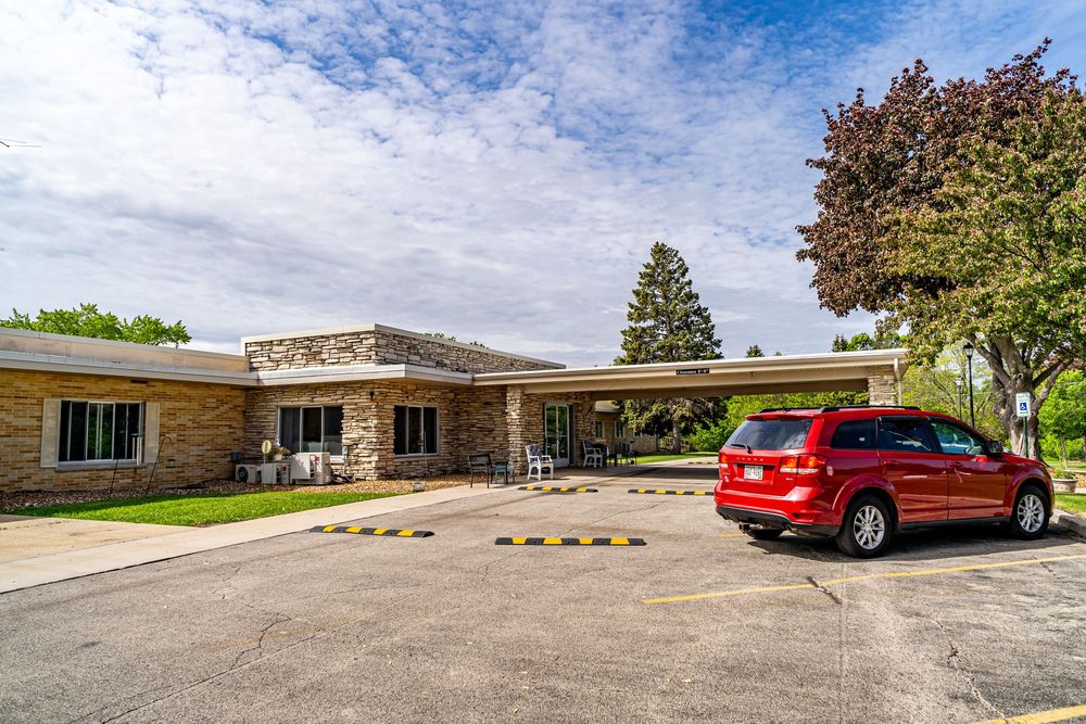 Single-story stone building with covered entrance, mature trees, and a red SUV parked in a spacious asphalt lot. real estate photography