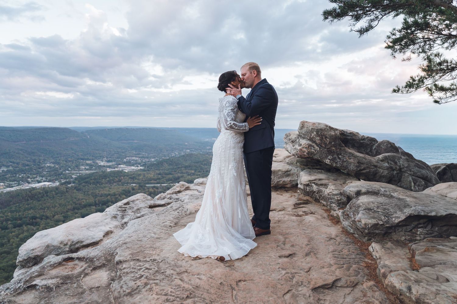 Bride and groom share a romantic kiss on a rocky mountain overlook with sweeping forest views under a cloudy sky.