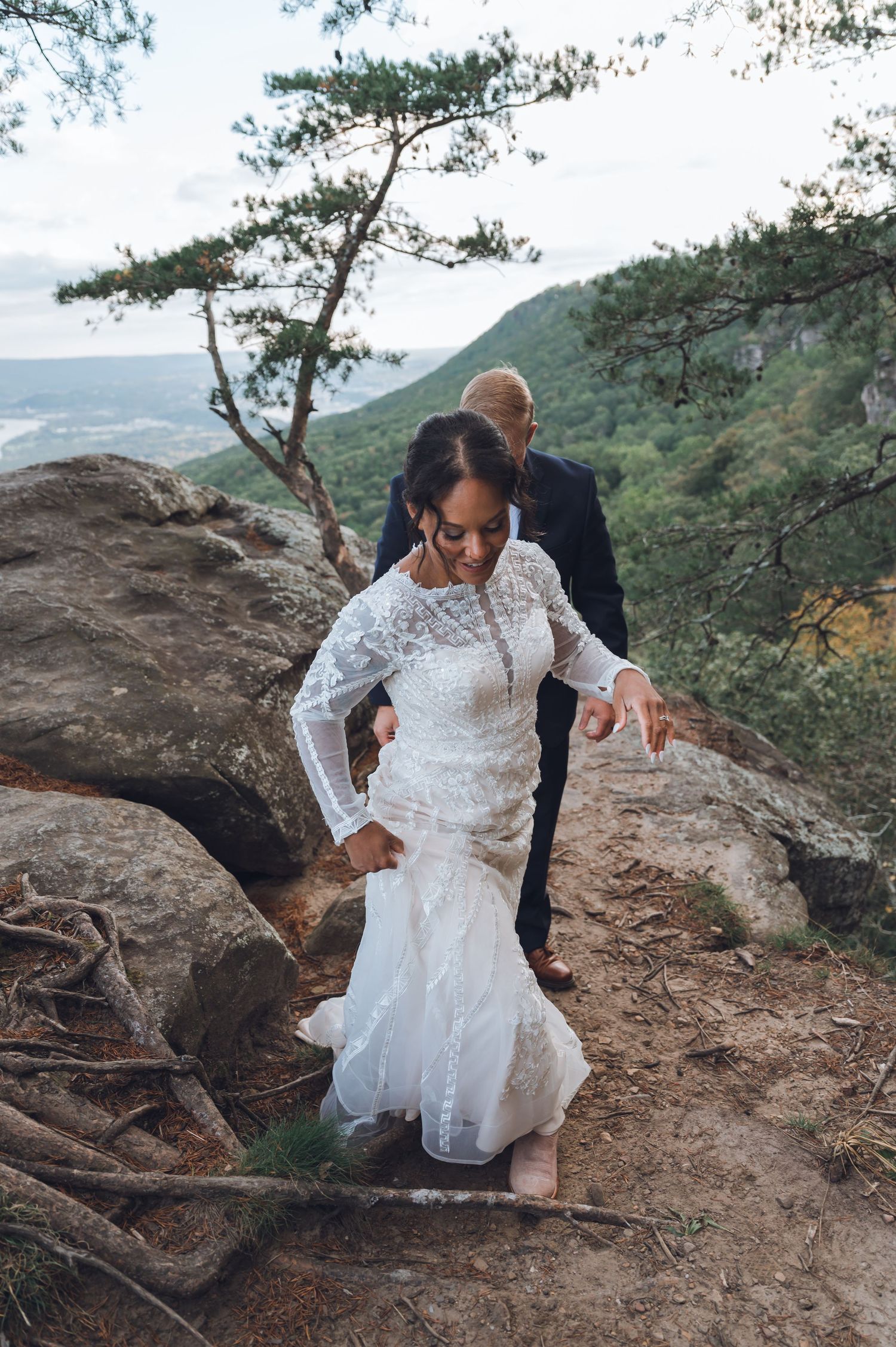 Bride in lace gown navigates rocky mountain trail with groom's assistance during outdoor wedding elopement.