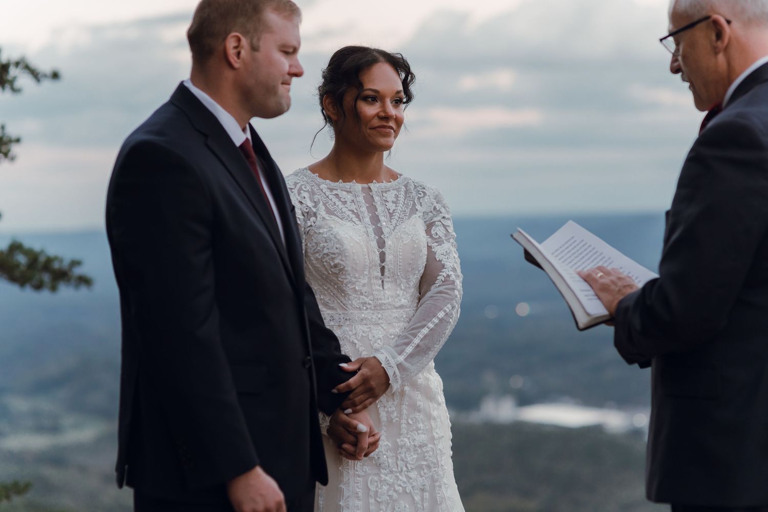 Couple exchanges wedding vows outdoors on a mountain overlook, officiant reads from book at scenic twilight ceremony.