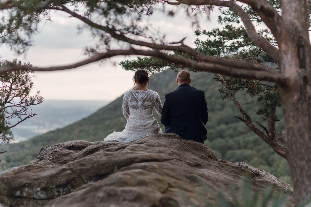 Bride and groom sitting on a rocky cliff overlooking a lush mountain landscape in their wedding attire.