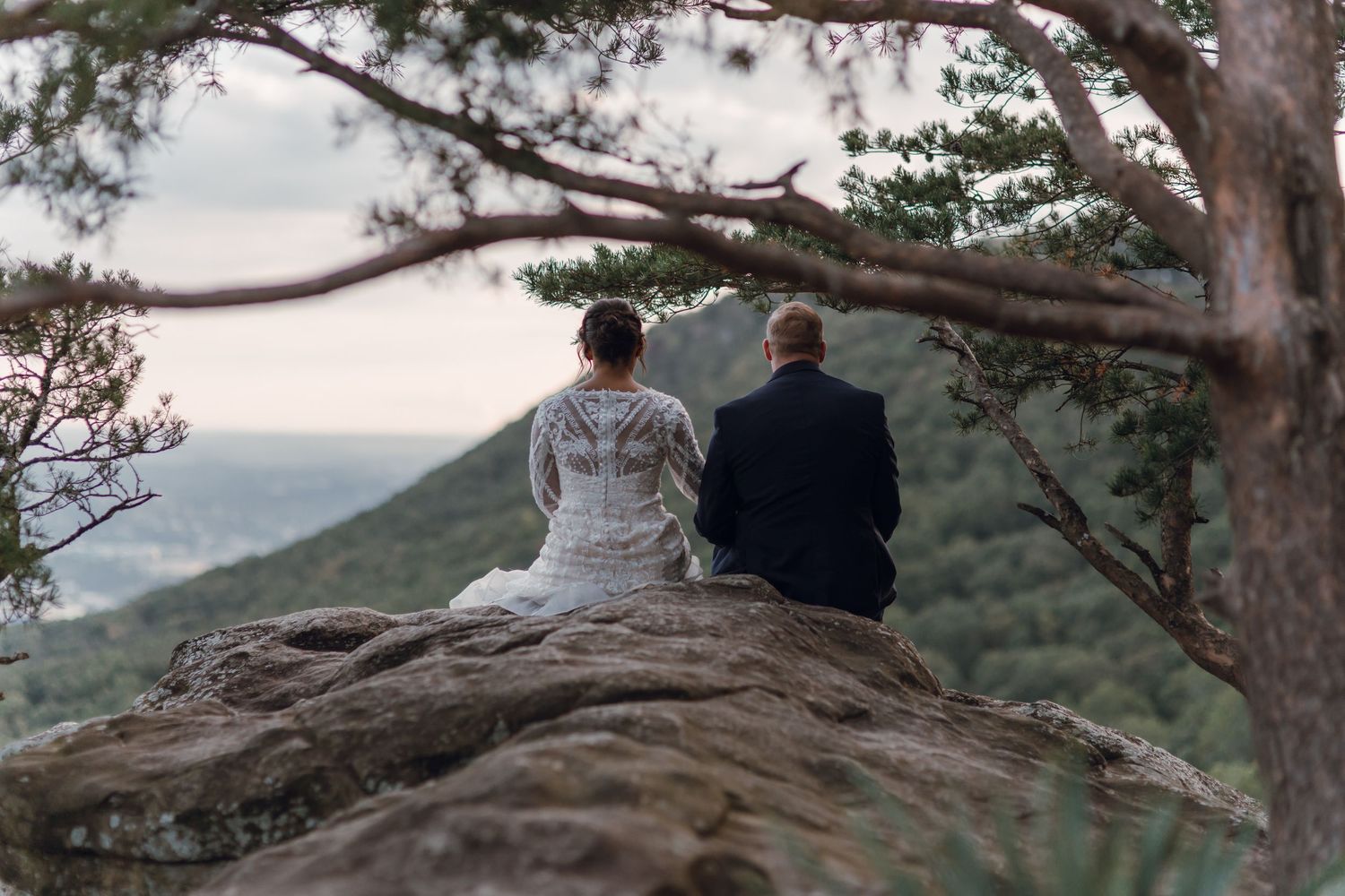 Bride and groom sitting on a rocky cliff overlooking a lush mountain landscape in their wedding attire.