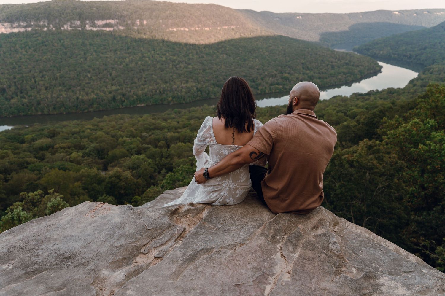 Couple sitting together on a rocky overlook enjoying a scenic view of a winding river through lush green forested hills.