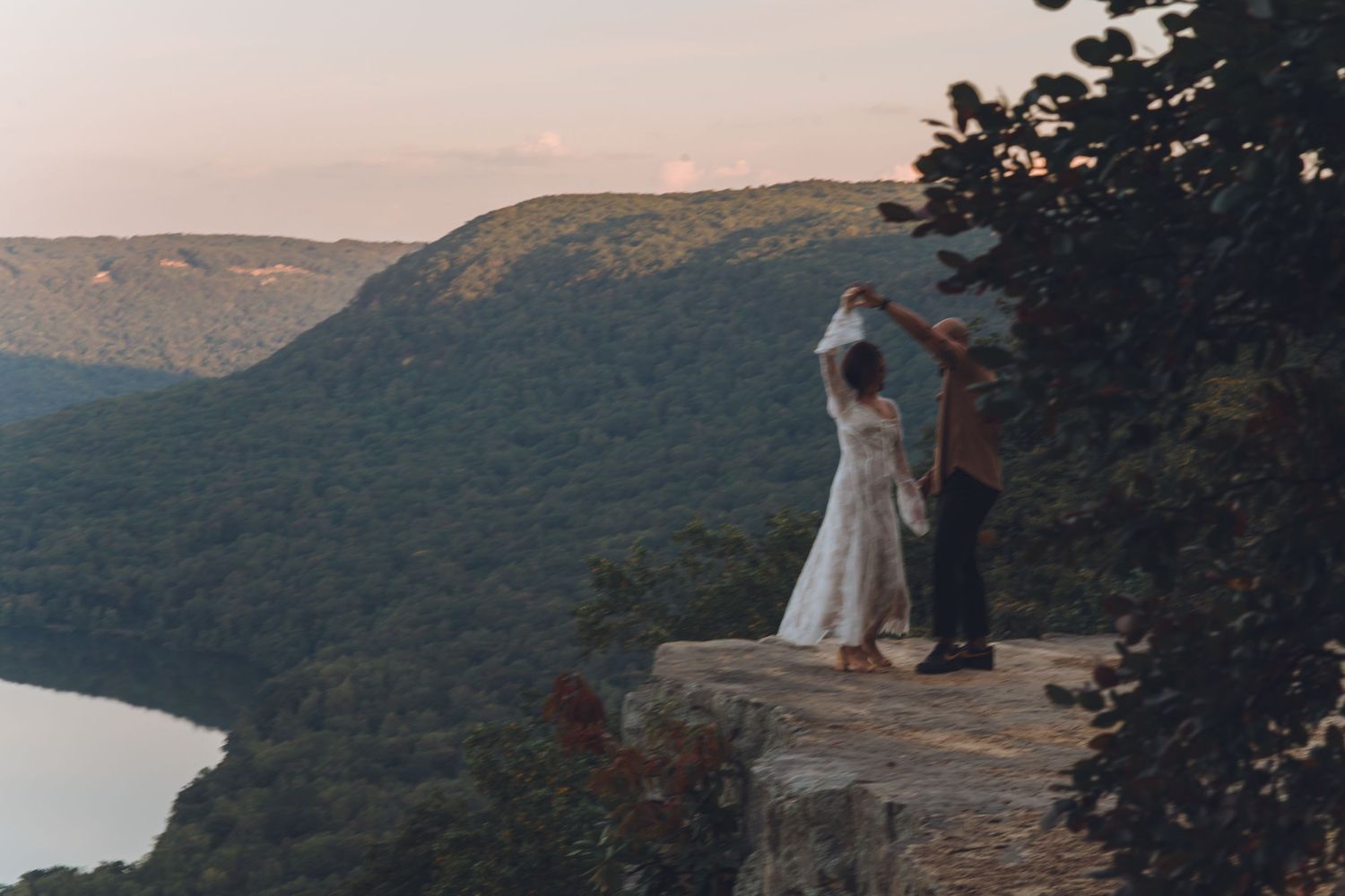Couple sharing a romantic dance on a rocky overlook with lush green mountains and river views at sunset.