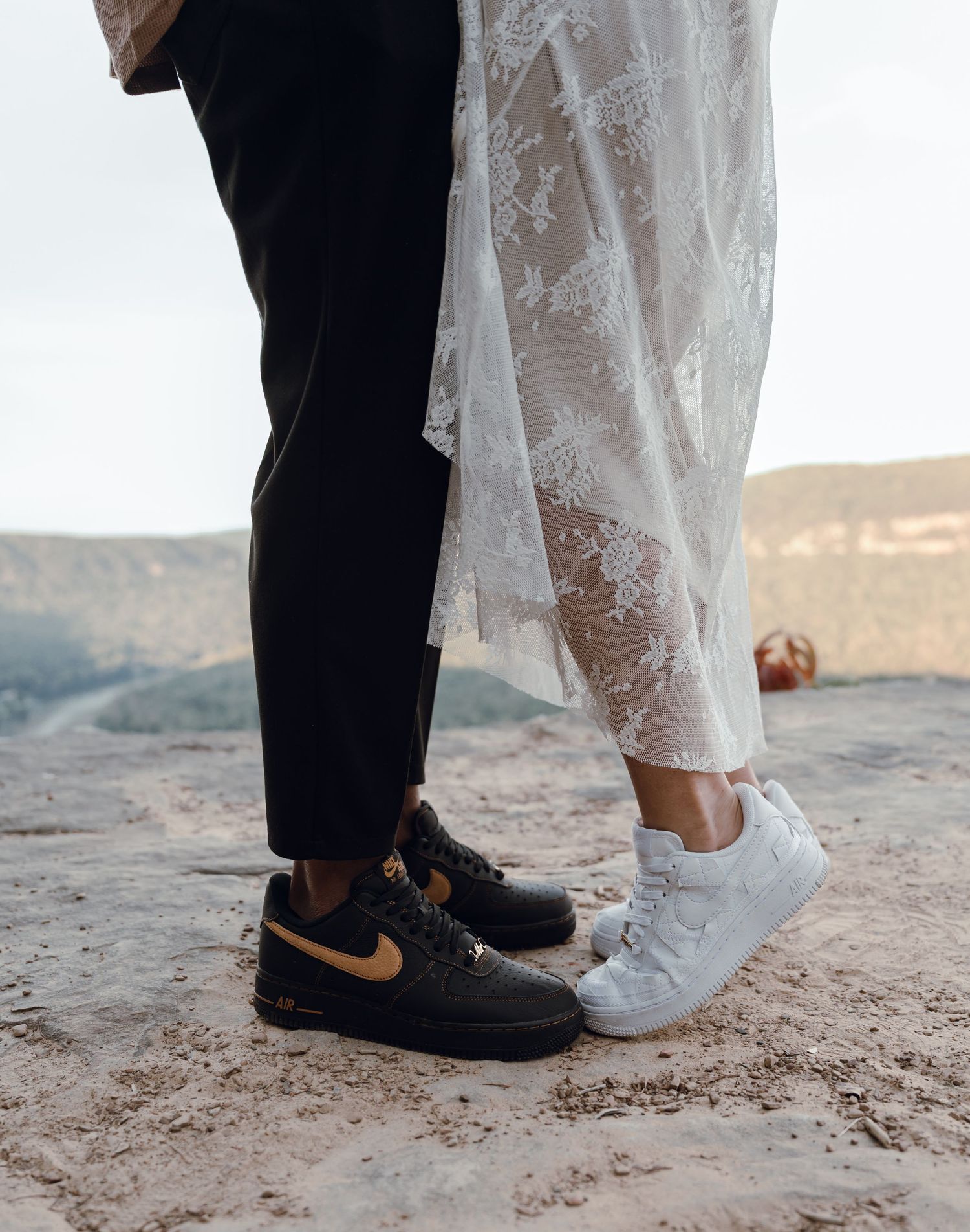 Bride and groom wearing matching Nike Air Force 1 sneakers, white and black with gold, standing on sandy terrain in wedding attire.