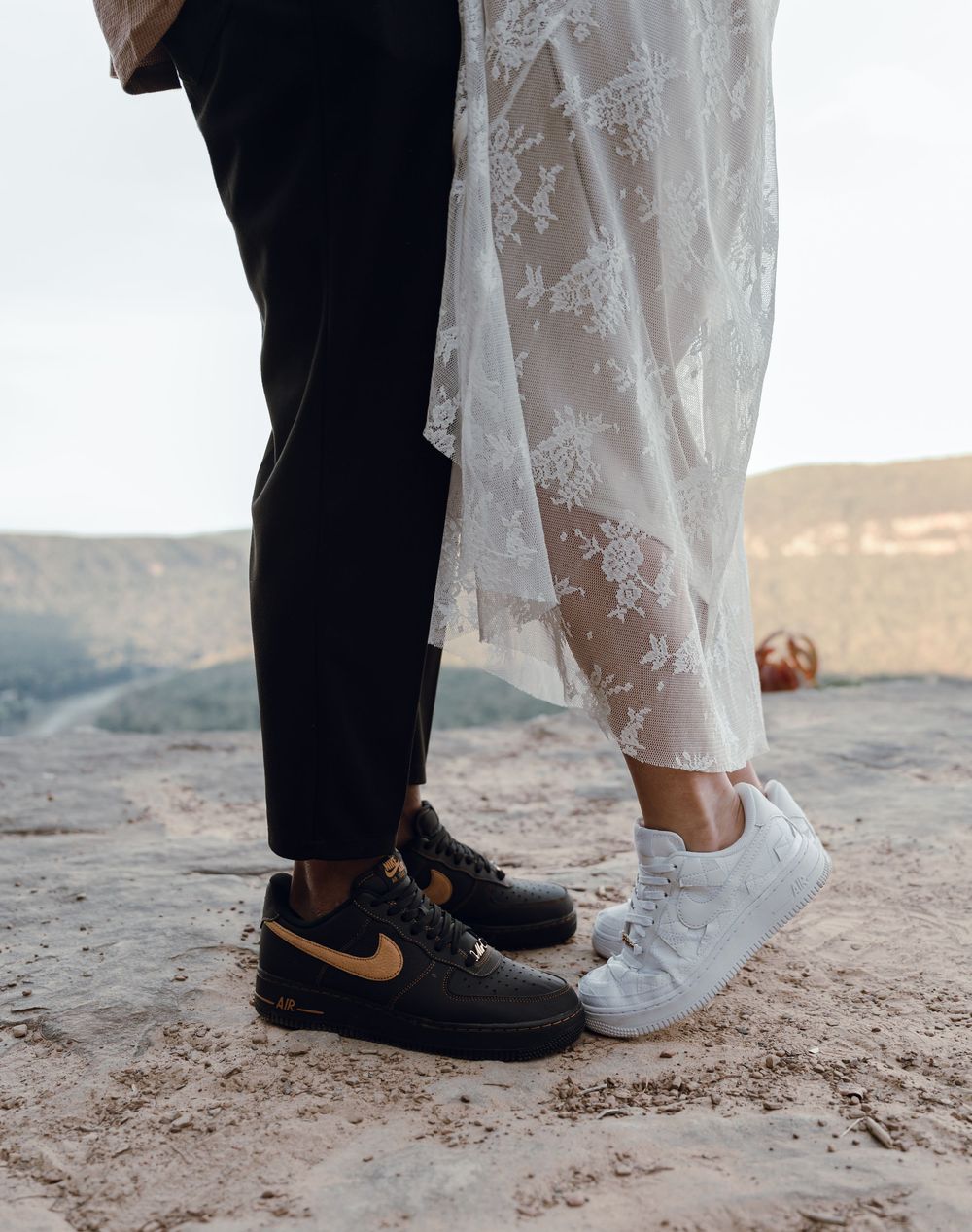 Bride and groom wearing matching Nike Air Force 1 sneakers, white and black with gold, standing on sandy terrain in wedding attire.