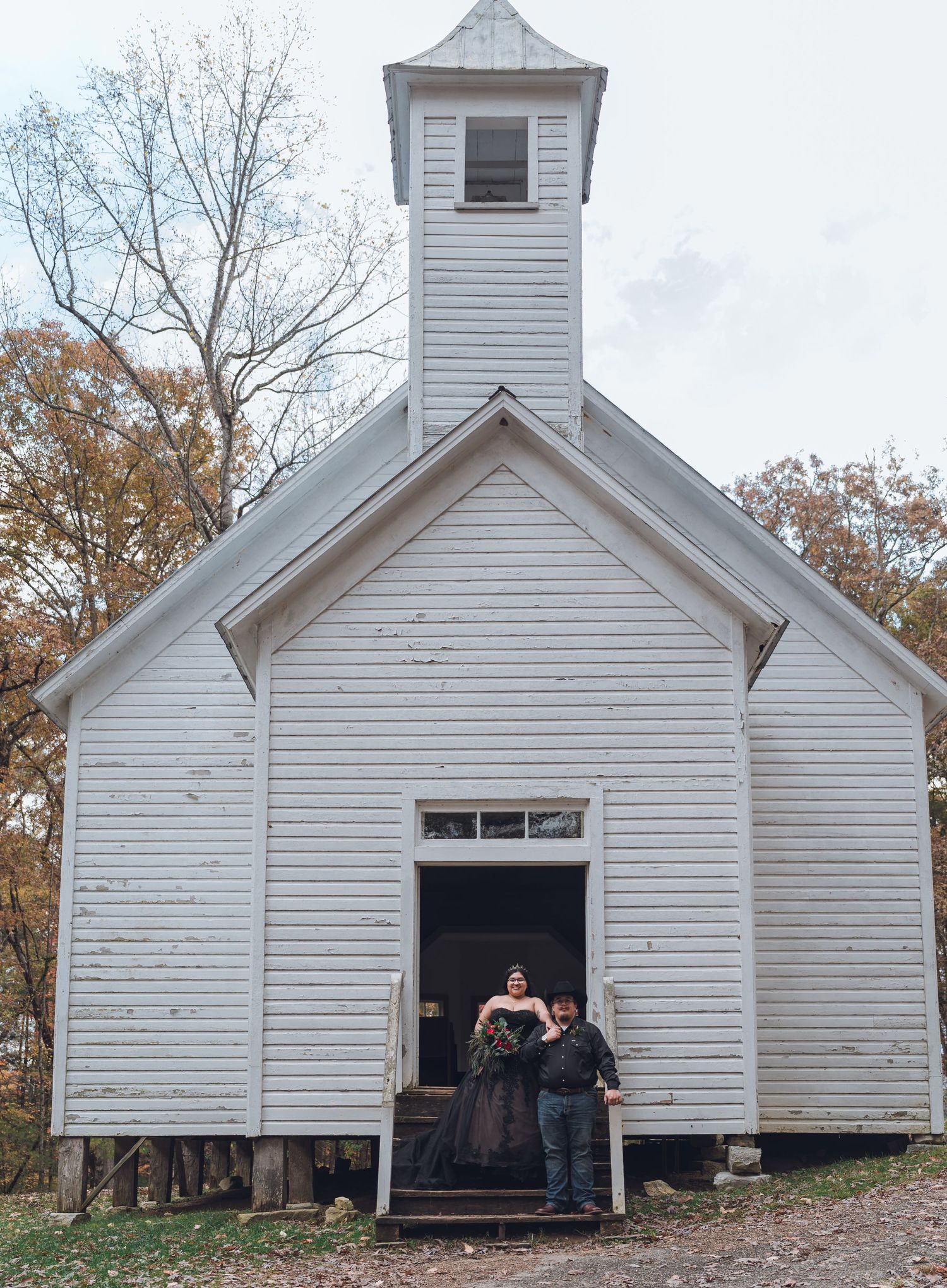 Couple stands at entrance of rustic white chapel church during fall outdoor wedding ceremony.