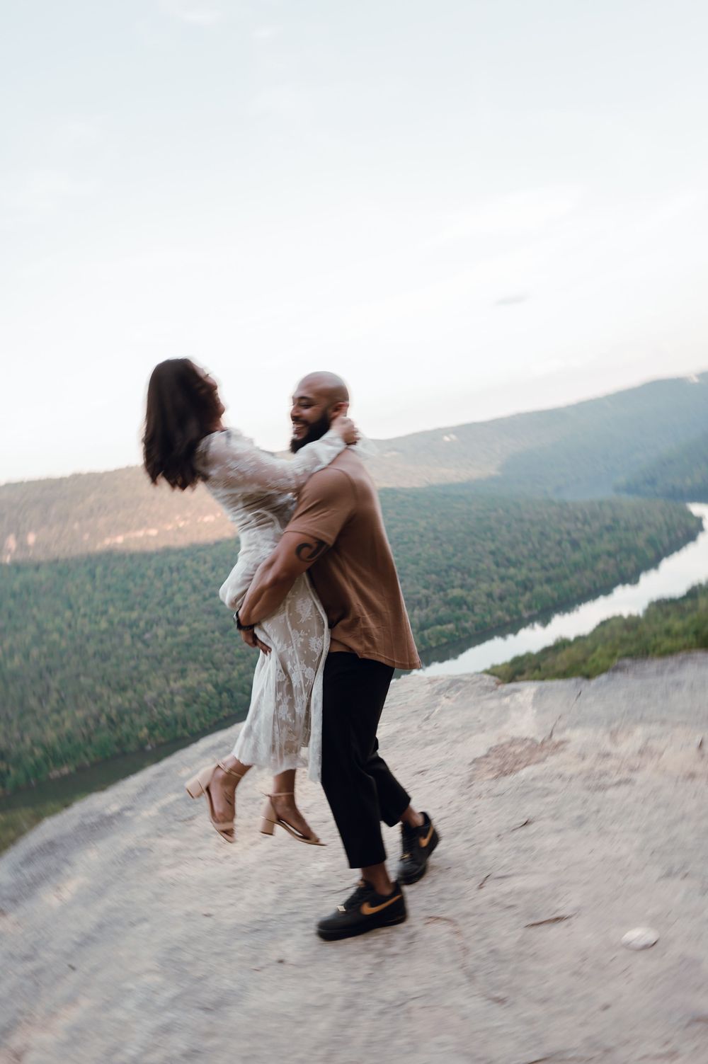 A man lifts a woman on a rocky cliff overlooking a lush forest and winding river at sunset.