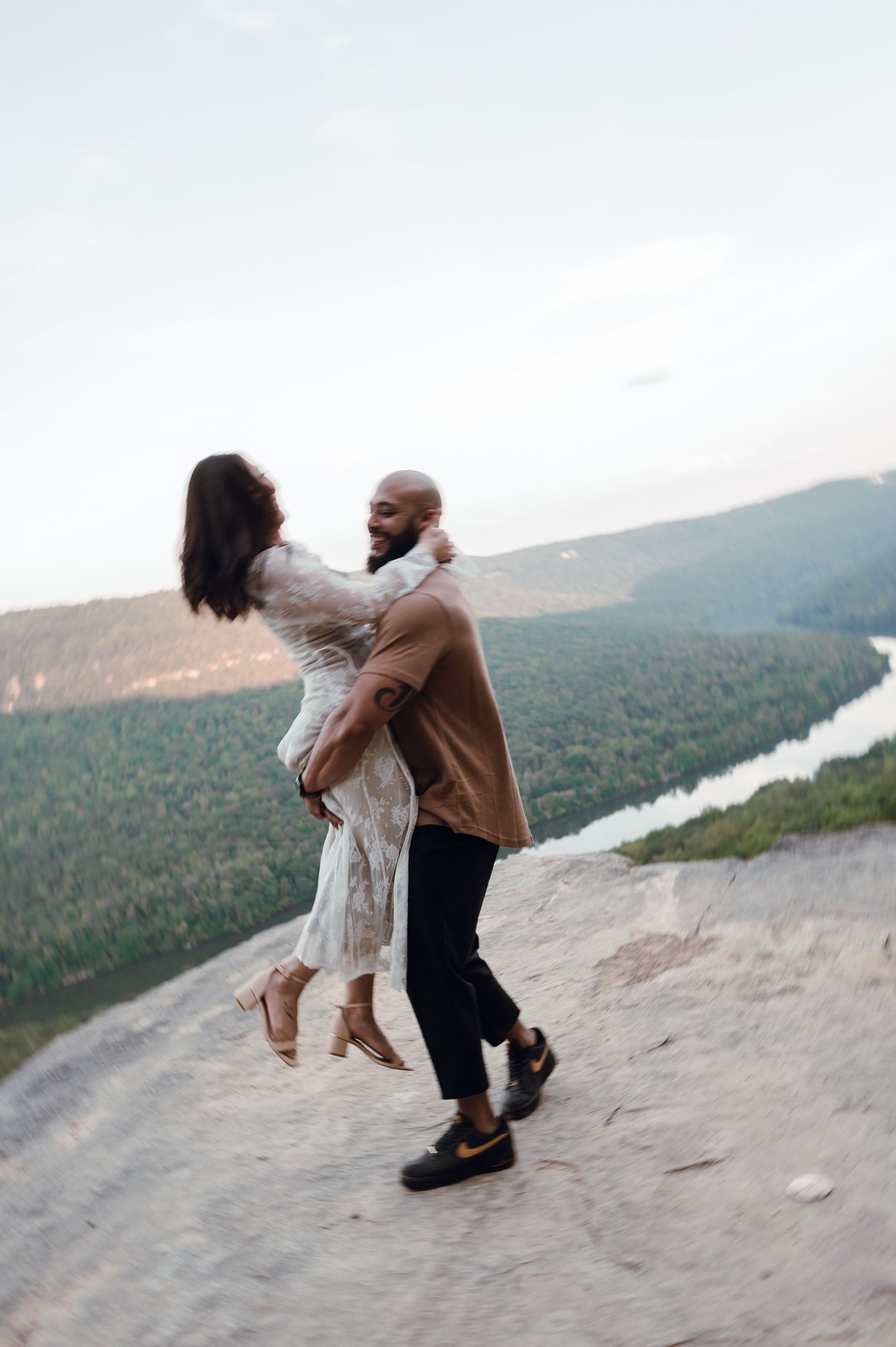 A man lifts a woman on a rocky cliff overlooking a lush forest and winding river at sunset.
