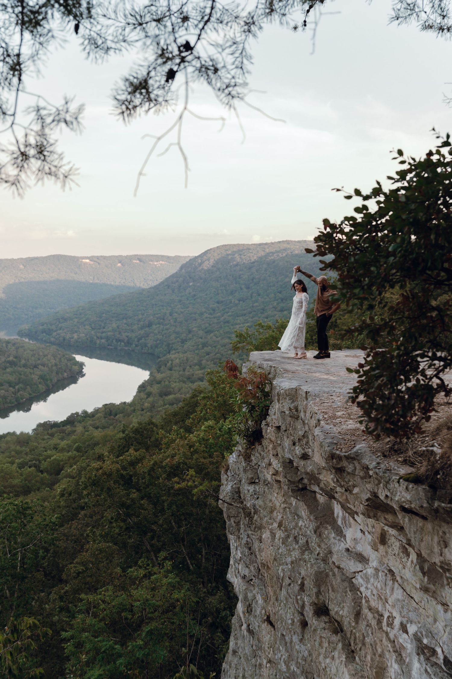 Couple in wedding attire dancing on a rocky cliff overlook with sweeping river valley and lush forest views below.