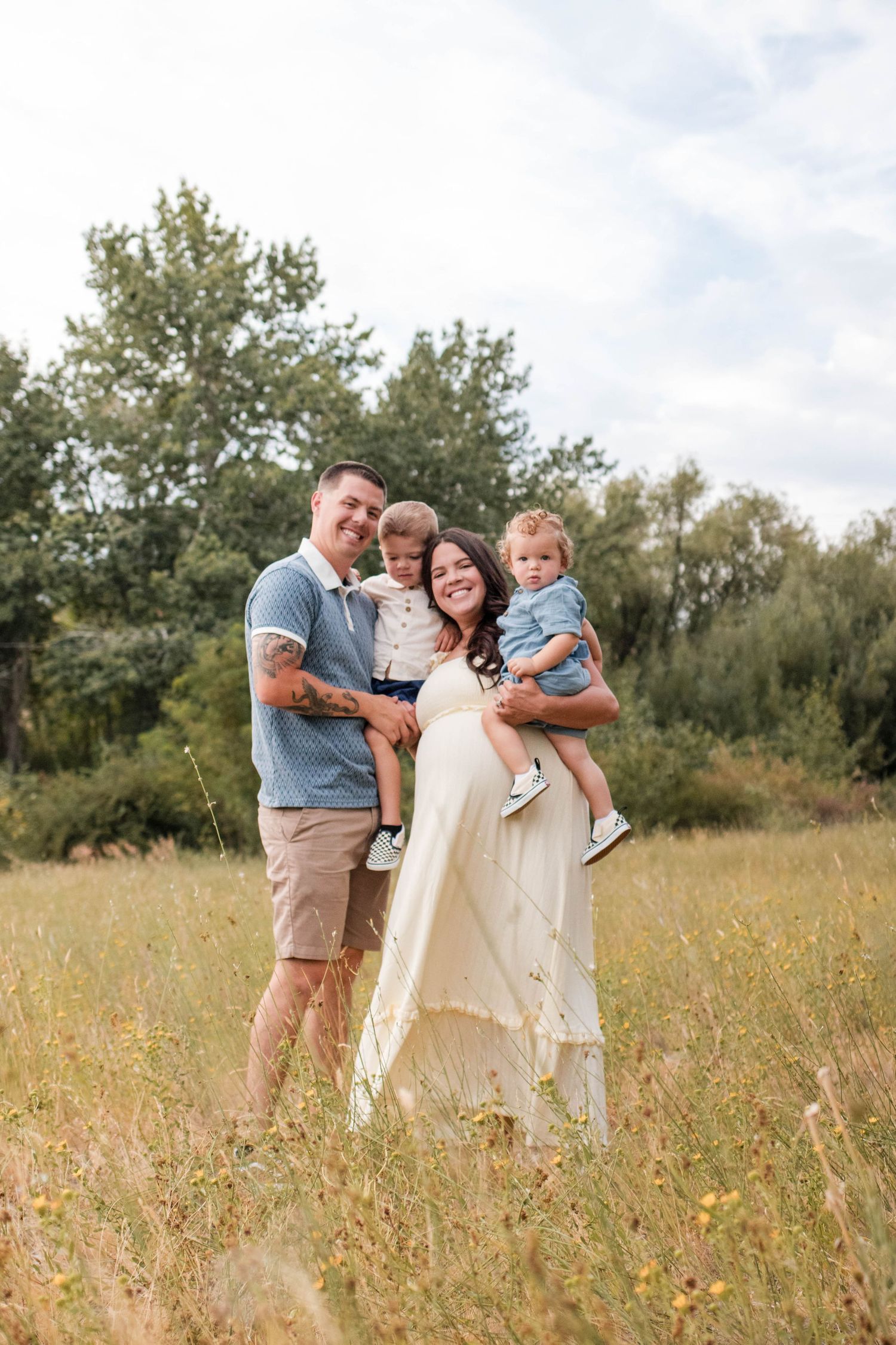 Happy family of four posing in a sunny meadow, parents holding two young children, surrounded by lush green trees.