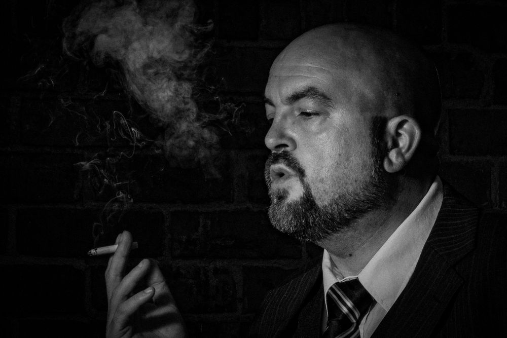 Black and white portrait of a bald bearded man in a suit smoking a cigarette against a brick wall.