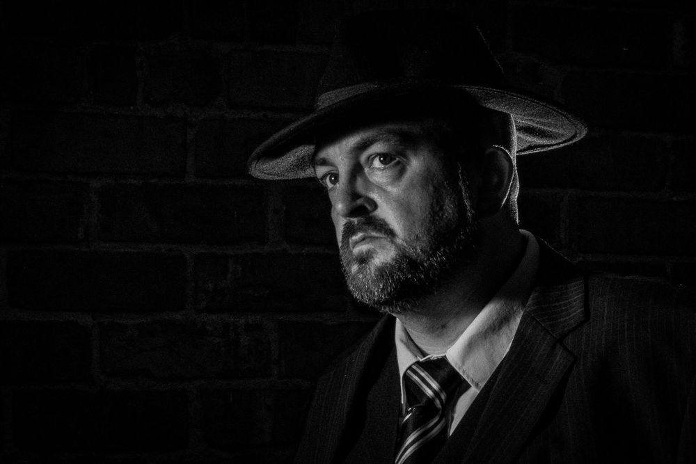 Black and white portrait of a bearded man wearing a fedora hat and suit against a dark brick wall.