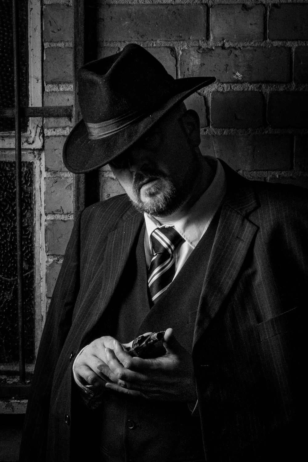 Black and white portrait of a man in a fedora hat and pinstripe suit holding a cigar against a brick wall.